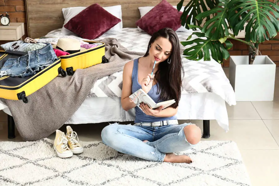 Young woman checking list of things while packing suitcase at home