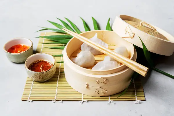Traditional chinese steamed dumplings Dim Sums HAR GOW in bamboo steamer with sauces and chopsticks atop tropical leaf on light surface with copy space.