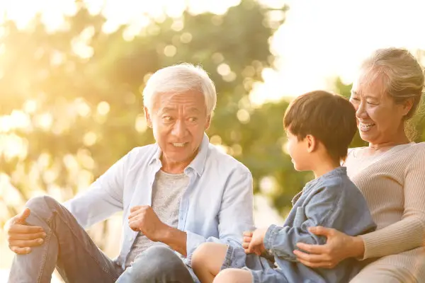 asian grandson, grandfather and grandmother sitting chatting on grass outdoors in park at dusk